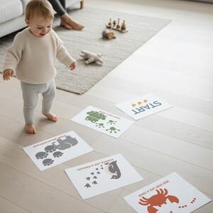Peut inclure: Un b&eacute;b&eacute; v&ecirc;tu d'un pull cr&egrave;me et d'un pantalon gris se tient debout sur un plancher en bois clair. Des cartes imprim&eacute;es avec des illustrations d'animaux et des instructions sont dispos&eacute;es sur le sol, dont une carte avec le mot "START".
