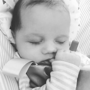 May include: Black and white image of a sleeping baby. The baby is nestled in a soft, patterned blanket, with closed eyes and a hand near the mouth. The baby is wearing a striped onesie.