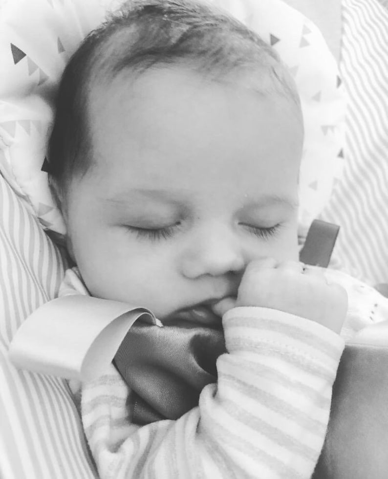 May include: Black and white image of a sleeping baby. The baby is nestled in a soft, patterned blanket, with closed eyes and a hand near the mouth. The baby is wearing a striped onesie.