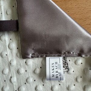 May include: A baby blanket with a gray satin border and a white, textured center. The blanket features a pattern of small black dots and giraffe illustrations. A tag reads "MAKES BY RACHII est. 2020".