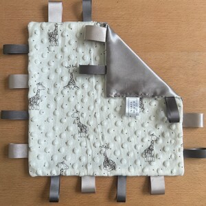 May include: A baby security blanket with a giraffe pattern. The ivory blanket has a textured surface and is trimmed with gray and tan ribbons. A corner is folded over, revealing a satin lining. The label reads "MAKES BY RACH".