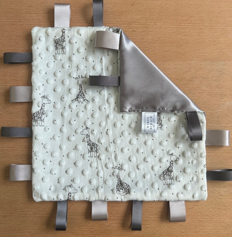 May include: A baby security blanket with a giraffe pattern. The ivory blanket has a textured surface and is trimmed with gray and tan ribbons. A corner is folded over, revealing a satin lining. The label reads "MAKES BY RACH".