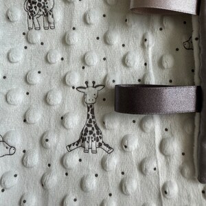 May include: A white baby blanket with a textured, dotted surface and black giraffe illustrations. The blanket has gray satin ribbons attached to the edge. The blanket is soft and plush, suitable for a baby.