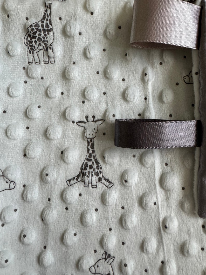 May include: A white baby blanket with a textured, dotted surface and black giraffe illustrations. The blanket has gray satin ribbons attached to the edge. The blanket is soft and plush, suitable for a baby.