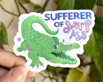 May include: A green alligator sticker with a purple and blue text overlay that reads "SUFFERER OF SWAMP ASS". The alligator has a toothy grin and a sparkly background.