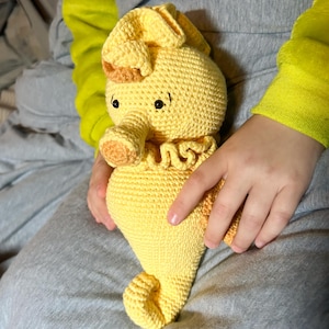 May include: A crocheted yellow seahorse toy with black eyes and a brown nose. The seahorse is being held by a person's hands.