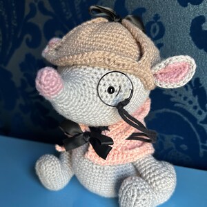 May include: A crocheted mouse wearing a tan newsboy cap, pink scarf, and a black bow tie. The mouse has a pink nose and ears, and a monocle. The toy is gray and white.