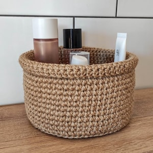 May include: A round, hand-crocheted basket made of natural jute fiber. The basket is filled with various cosmetic products. The basket is sitting on a wooden surface, with a white tiled wall in the background.