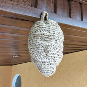 Op de afbeelding: Een beige gehäkelde hangende tas met een lus aan de bovenkant, hangend aan een houten plafond.