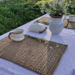 Puede incluir: Una mesa rústica al aire libre con un mantel blanco con lunares grises, adornada con manteles individuales marrones tejidos, una mantequera y una jarra de cerámica llena de flores blancas. Una taza y una taza también están sobre la mesa.