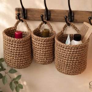 Puede incluir: Tres cestas cilíndricas de yute tejidas a mano con asas integradas, colgadas de un perchero de madera con cuatro ganchos de metal negro. Las cestas contienen varios artículos de aseo personal, incluyendo botellas y un peine de madera.