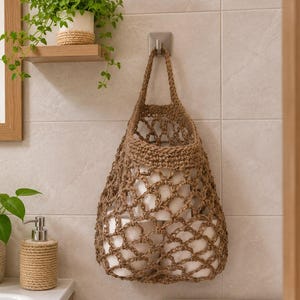 Pode incluir: Uma bolsa de armazenamento suspensa de crochê marrom, feita de corda grossa com um padrão de rede de malha aberta. A bolsa está suspensa em um gancho de parede e contém vários rolos de papel higiênico brancos, exibindo um design funcional.