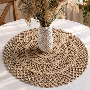 May include: A round, tan crocheted doily with an intricate openwork pattern sits on a white tablecloth. A white, textured, leaf-shaped ceramic vase holds a tall arrangement of dried, feathery beige grasses with small green accents.
