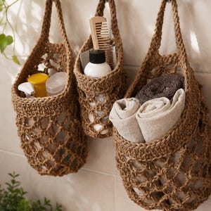 May include: Three brown jute hanging baskets suspended from a silver metal wall rack. The baskets feature an open net weave design and contain various bathroom items including a wooden comb, a white bottle, and folded beige and brown towels.
