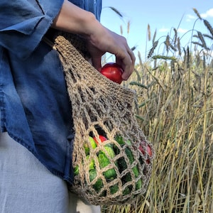 Peut inclure: Une personne portant une chemise en jean bleu et un pantalon blanc tient un sac en filet brun rempli de concombres verts et de tomates rouges dans un champ d'herbe sèche et haute.