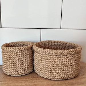 Puede incluir: Dos cestas redondas de ganchillo en color beige natural. Las cestas están hechas de hilo grueso y tienen un aspecto texturizado. Están sobre una superficie de madera contra un fondo de baldosas blancas.