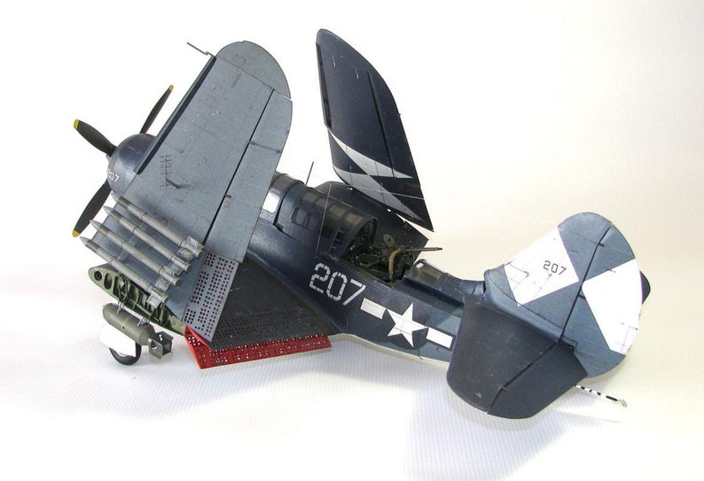 May include: A blue and white model airplane with the number 207 on the side. The plane has its wings folded back and is sitting on a white surface.