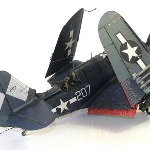 May include: A blue and white model airplane with the number 207 on the side. The plane has its wings folded back and is sitting on a white surface. The plane has a star on the tail and a red and white stripe on the bottom.