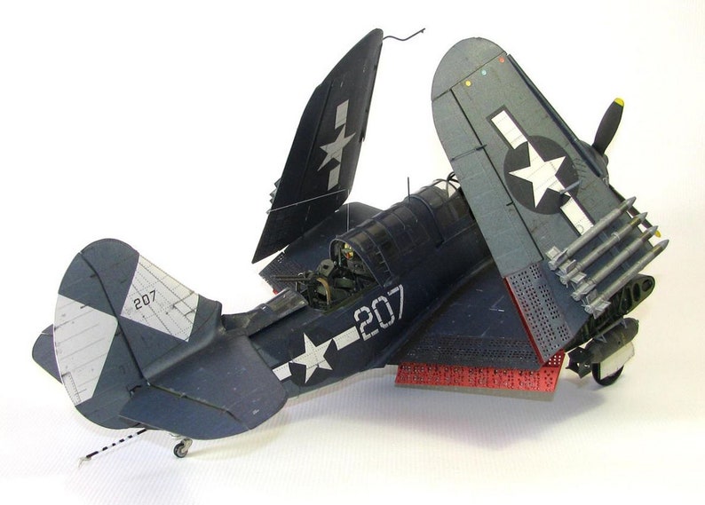 May include: A blue and white model airplane with the number 207 on the side. The plane has its wings folded back and is sitting on a white surface. The plane has a star on the tail and a red and white stripe on the bottom.