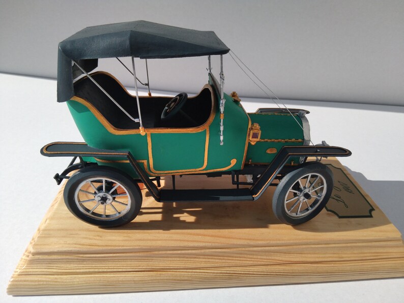 Puede incluir: Un coche modelo verde y dorado con una capota de tela negra. El coche est&aacute; sobre una base de madera con una placa que dice "1907".