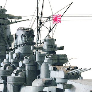May include: A close-up of a grey model of a battleship with two red Japanese flags flying from the masts. The ship has multiple gun turrets and other details.