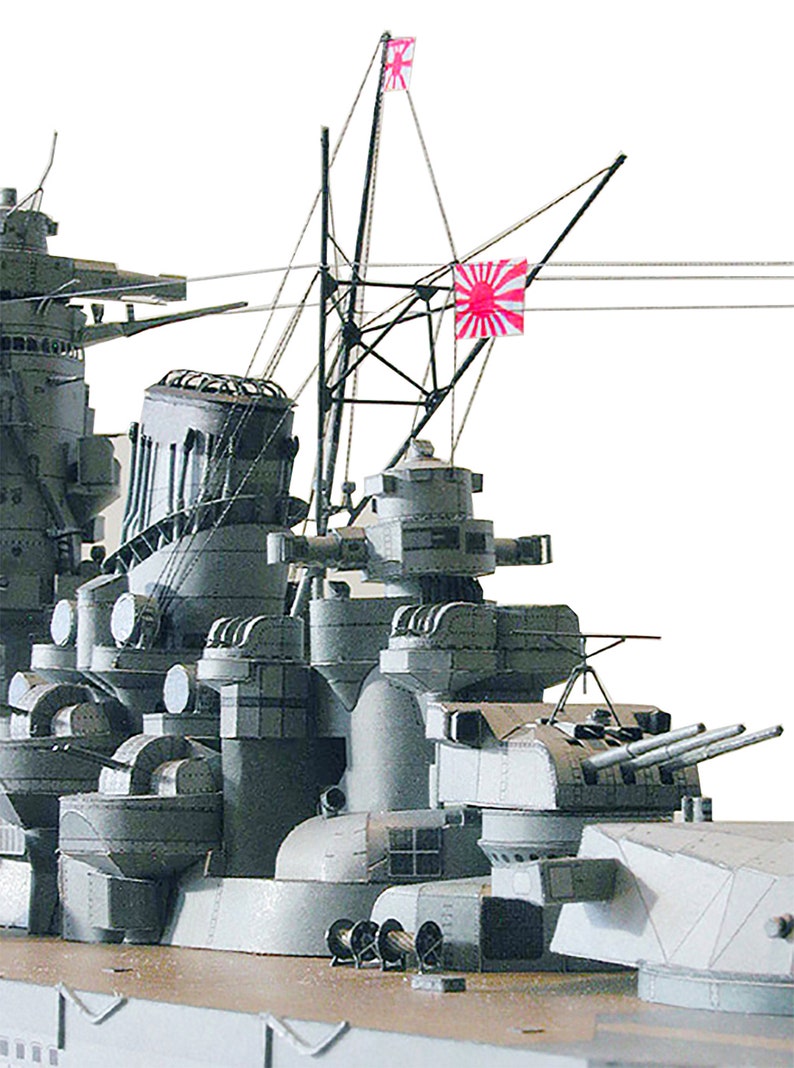 May include: A close-up of a grey model of a battleship with two red Japanese flags flying from the masts. The ship has multiple gun turrets and other details.
