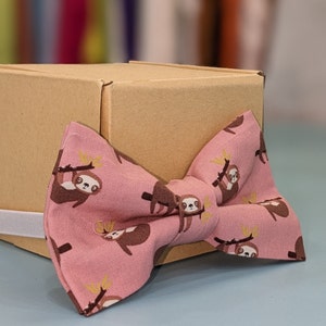 May include: A pink bow tie with a pattern of brown sloths hanging from branches. The bow tie is on top of a brown cardboard box.