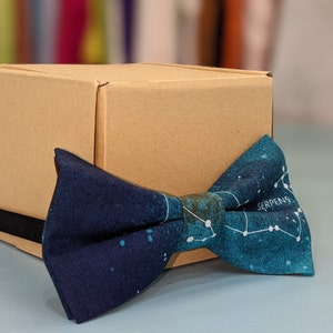 Peut inclure: Un nœud papillon bleu foncé et turquoise avec un motif de constellation et le mot "Serpens" imprimé dessus. Le nœud papillon est posé sur un carton brun.