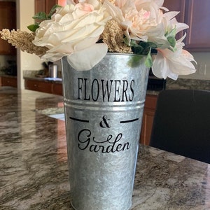 Może przedstawiać: Galwanizowany metalowy wazon z czarno-białym wzorem, na którym widnieje napis "Flowers & Garden". Wazon jest wypełniony bukietem białych i różowych kwiatów.
