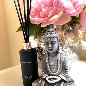 K&ouml;nnte beinhalten: Eine schwarze Diffuserflasche mit schwarzen Ruten und dem Text "Federico Mahora Pure Home" steht auf einer wei&szlig;en Oberfl&auml;che neben einer grauen Steinbuddha-Statue. Der Buddha sitzt in meditativer Haltung mit den H&auml;nden im Scho&szlig;. Eine Vase mit rosa Pfingstrosen steht im Hintergrund.