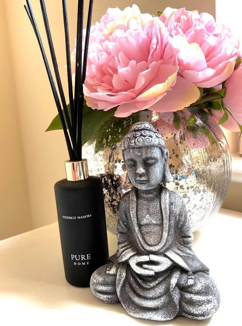 K&ouml;nnte beinhalten: Eine schwarze Diffuserflasche mit schwarzen Ruten und dem Text "Federico Mahora Pure Home" steht auf einer wei&szlig;en Oberfl&auml;che neben einer grauen Steinbuddha-Statue. Der Buddha sitzt in meditativer Haltung mit den H&auml;nden im Scho&szlig;. Eine Vase mit rosa Pfingstrosen steht im Hintergrund.