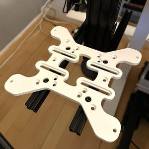 May include: A white, geometric bracket with multiple holes and slots, mounted on black rails. The bracket has a symmetrical design with rounded edges and is secured with black screws. The background includes a black structure and wooden flooring.