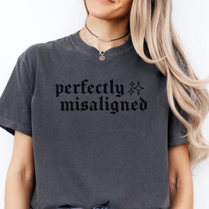 May include: A charcoal gray short-sleeve t-shirt featuring the black text "perfectly misaligned" printed across the chest in a gothic-style font, accompanied by two small four-pointed star icons positioned to the right of the lettering.