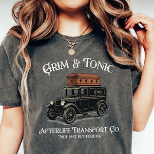May include: Pepper-colored t-shirt featuring a vintage hearse graphic with a coffin on top. The text reads "Grim & Tonic" above the hearse and "Afterlife Transport Co." below, with the tagline "Not fast, but forever."