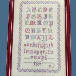 May include: A framed cross-stitch of the alphabet in different colors. The letters are in a traditional font and are surrounded by a border of pink and blue zigzags. The piece is dated 1986 and signed "M.C."