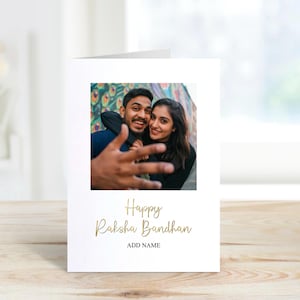 Puede incluir: Una tarjeta de felicitación blanca de Raksha Bandhan con una fotografía de una pareja sonriente. La tarjeta presenta una escritura dorada que dice "Happy Raksha Bandhan" y "ADD NAME" debajo. La tarjeta se muestra sobre una superficie de madera clara.