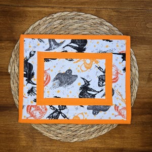 May include: A rectangular quilted fabric mat featuring a white background with a repeating Halloween pattern of black ghosts, witches, and orange pumpkins, accented by orange fabric borders and a central orange rectangular frame detail.