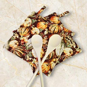 May include: Two cream-coloured wooden spoons rest on top of two square quilted pot holders. The pot holders feature a repeating pattern of pumpkins in shades of orange, yellow, and green on a dark brown background.