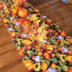 May include: A black table runner with a vibrant autumn pattern featuring pumpkins, sunflowers, and fall leaves in orange, yellow, and green. Decorative pumpkins and leaves are arranged on top.