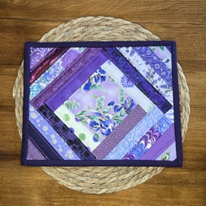 May include: A square quilted potholder featuring a diagonal log cabin pattern in various shades of purple, lavender, and violet. The centre square displays a floral print with blue and purple irises, surrounded by strips of fabric with floral patterns.