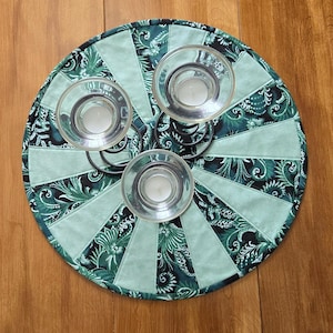 May include: A circular, quilted candle holder with three glass candle cups. The holder has a fan design with alternating sections of teal and patterned fabric in green and black. The metal candle holder is black.