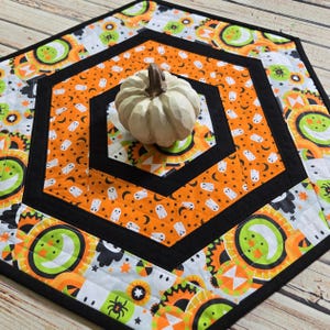 May include: A hexagonal quilted table mat featuring a Halloween-themed pattern of white ghosts, black spiders, and orange moons on a white background, bordered by black fabric strips, with a small, off-white, carved decorative pumpkin in the centre.