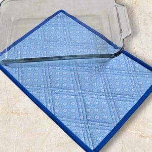 Peut inclure: Un plat à four en verre transparent repose sur un dessous de plat bleu matelassé. Le dessous de plat présente un motif floral et une bordure bleu foncé. Il est conçu pour protéger les surfaces de la chaleur.