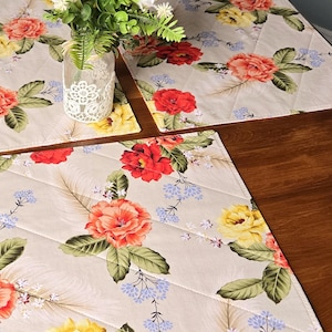 Peut inclure: Sets de sets de table matelassés à motifs floraux, avec un fond crème, des roses rouges, jaunes et oranges, des feuilles vertes et de petites fleurs bleues. Un vase en verre transparent avec de la dentelle blanche contient des fleurs blanches et vertes.