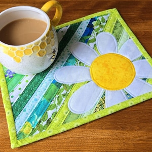 May include: A white ceramic mug with a yellow honeycomb pattern and a bee illustration, filled with coffee, sits on a square fabric coaster. The coaster features a large white daisy with a yellow centre over diagonal green and blue fabric strips.