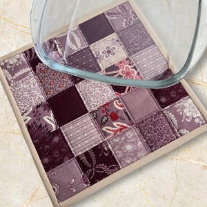 May include: A patchwork quilted hot pad in shades of purple, burgundy, and cream, with a beige border. The square design features various floral and patterned fabrics. A clear glass baking dish rests on top.