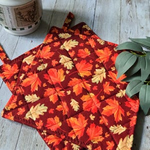 May include: A square quilted potholder featuring a deep burgundy background with a repeating pattern of autumn leaves in shades of orange, yellow, and light tan. The fabric is stitched in a diamond grid pattern and includes a small fabric hanging loop.