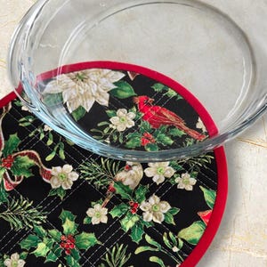 Könnte beinhalten: Eine klare Glasbackform liegt auf einem gesteppten, schwarzen, weihnachtlichen Topflappen. Der Topflappen zeigt einen roten Kardinal, weiße Blumen, Stechpalmen und einen roten Rand. Der Topflappen hat einen Durchmesser von etwa 20 cm.