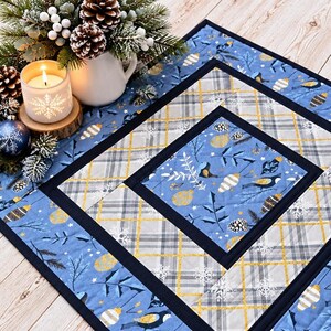 May include: A quilted table runner featuring a blue, grey, and gold pattern with holiday ornaments, pine branches, and plaid sections. The fabric is bordered by dark navy strips and displays a repeating design of birds and snowflakes.