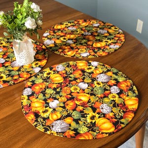 May include: Two round fabric placemats featuring a black background with a vibrant pattern of orange pumpkins, yellow sunflowers, green leaves, and white decorative pumpkins. The placemats are quilted with visible circular stitching.